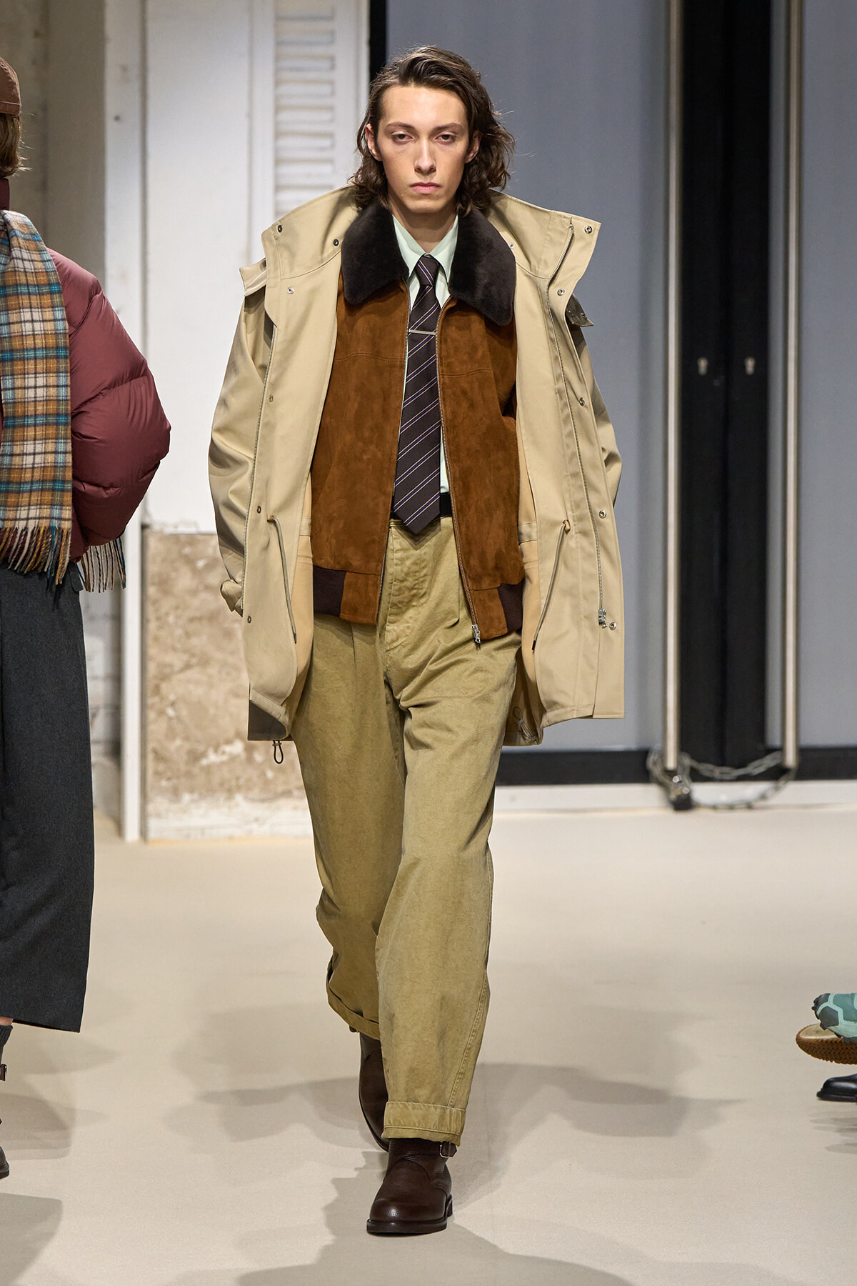Male model on a runway wearing a beige trench coat over a brown suede vest, striped tie, light green shirt, and khaki pants with brown boots.