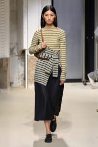Model walking a fashion runway in a beige striped knit top with a matching wrapped belt over a black midi skirt, accessorized with a brown shoulder bag and black loafers.