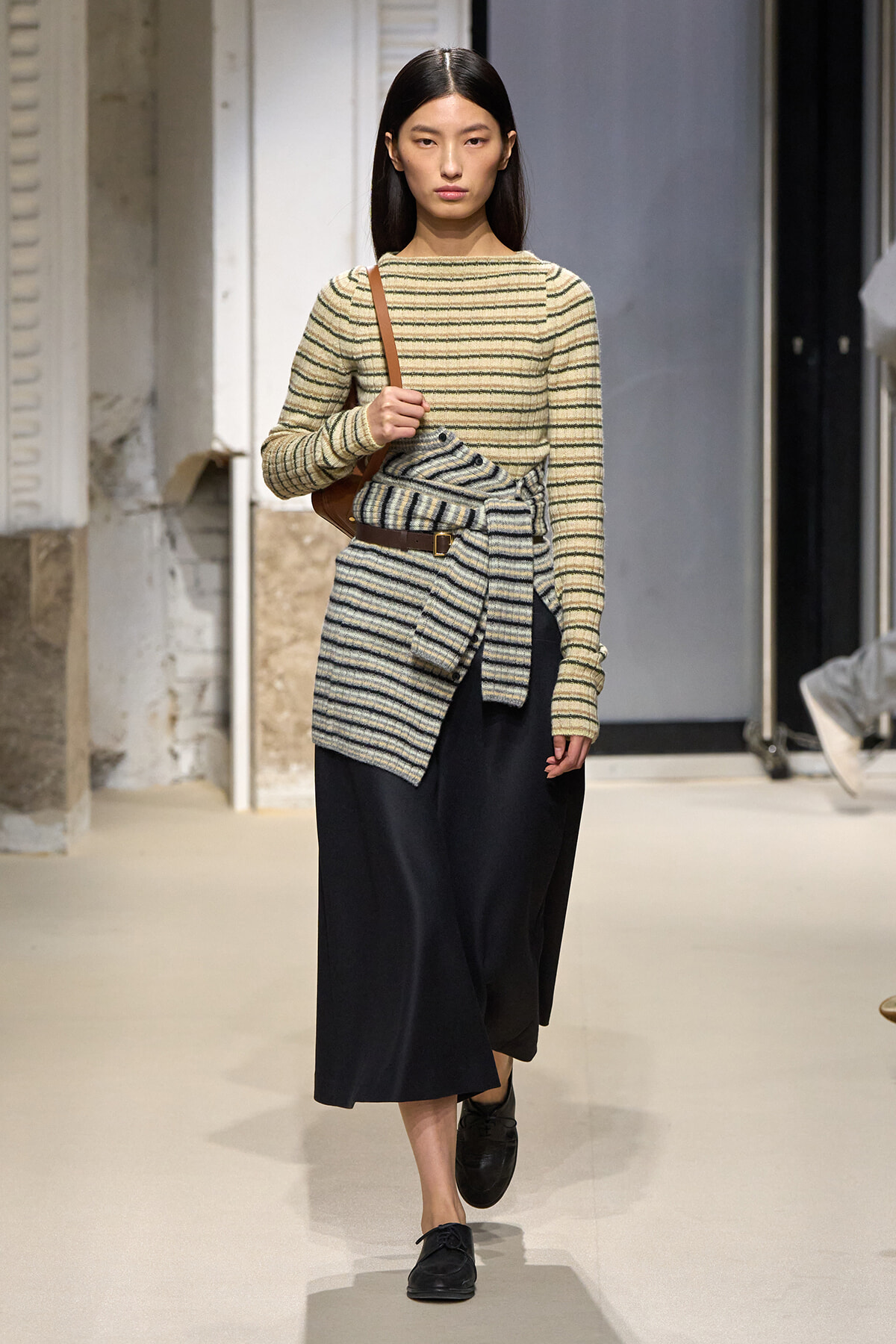 Model walking a fashion runway in a beige striped knit top with a matching wrapped belt over a black midi skirt, accessorized with a brown shoulder bag and black loafers.