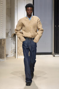 Male model walking a runway in a tan chunky knit cardigan over a blue collared shirt with dark denim jeans and black shoes.