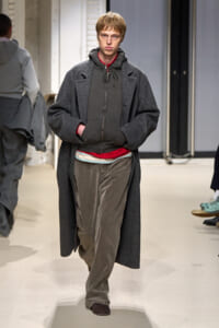 Male model on a runway wearing a long dark gray overcoat over a charcoal hoodie with a red shirt peeking at the hem, loose brown pants, and brown shoes.