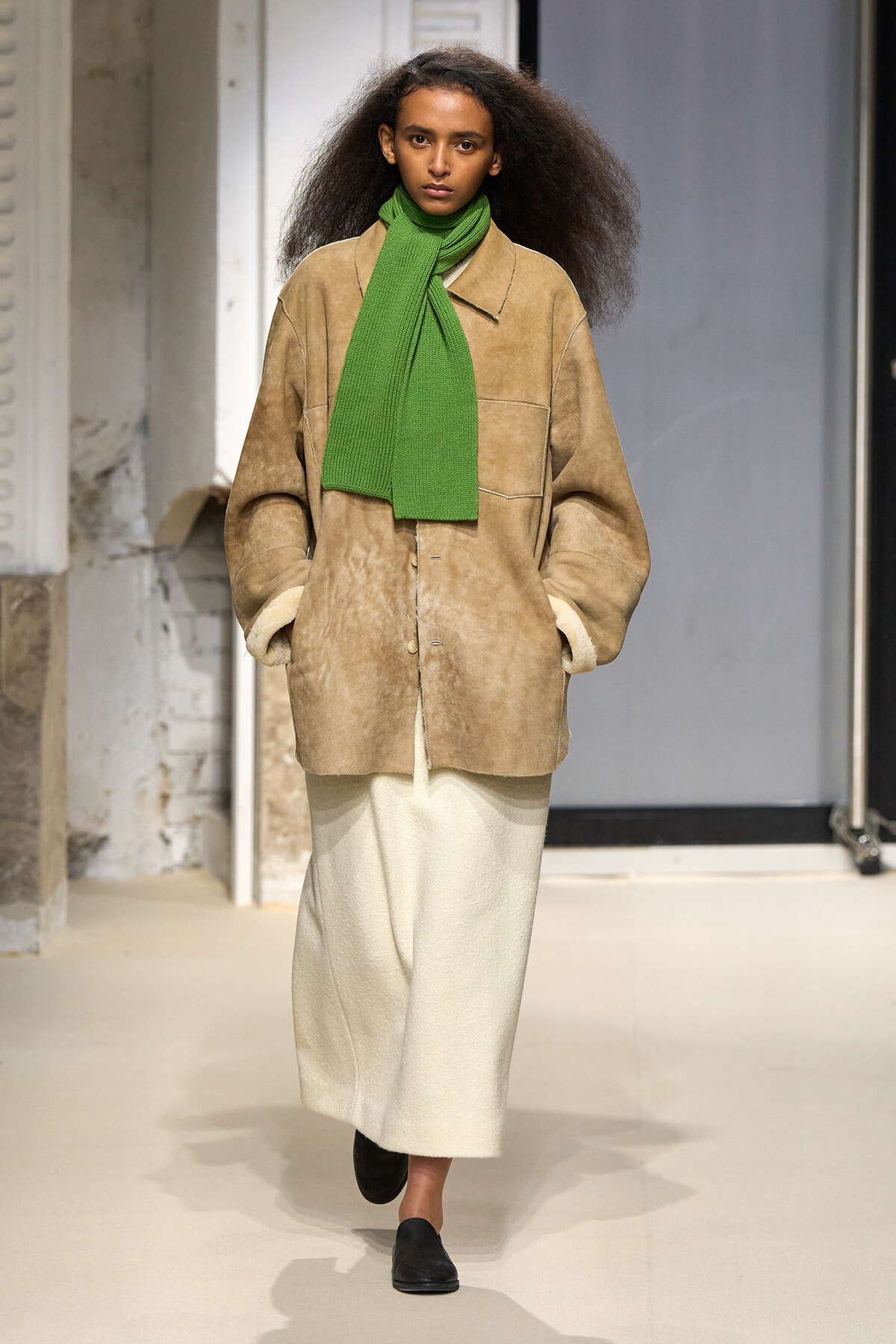 Runway model wearing a tan oversized suede coat, white midi skirt, black shoes, and a bright green scarf.