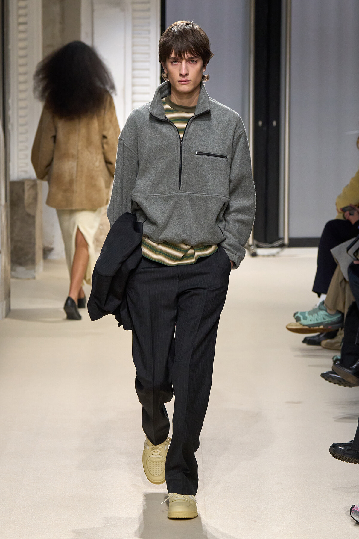 Male model walking a runway in a gray quarter-zip fleece, striped shirt underneath, black pinstripe trousers, beige sneakers, and a dark jacket draped over arm.