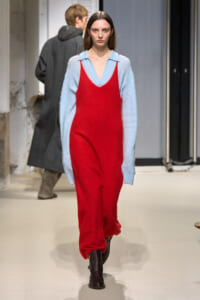 Model walking the runway in a bright red loose jumpsuit over a light blue V-neck sweater, with dark boots.