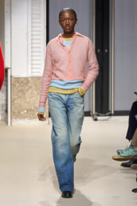 Model walking the runway in a pastel pink cardigan over layered blue tops and wide-leg jeans.