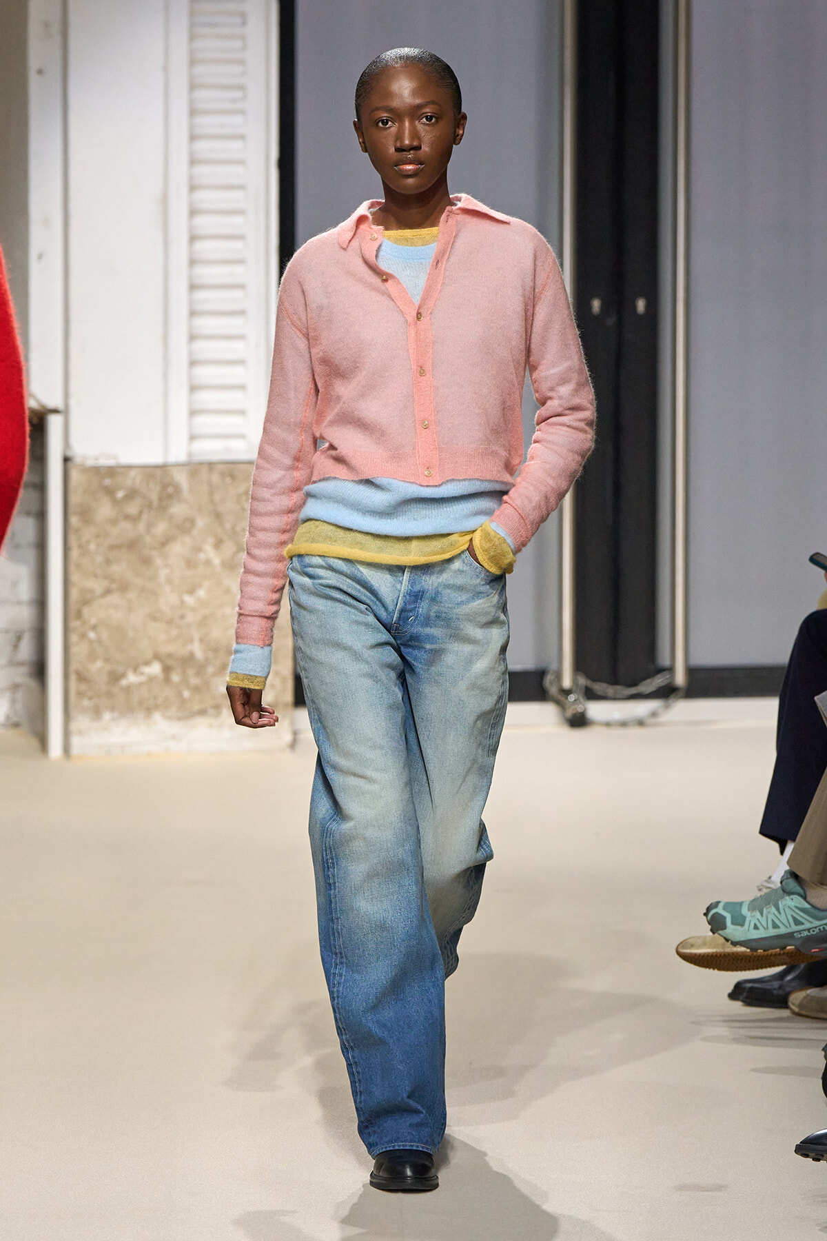 Model walking the runway in a pastel pink cardigan over layered blue tops and wide-leg jeans.
