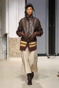 Runway model wearing a brown leather jacket, cream knit layer peeking at the hem, beige wide-leg pants, and dark boots with a black cap.