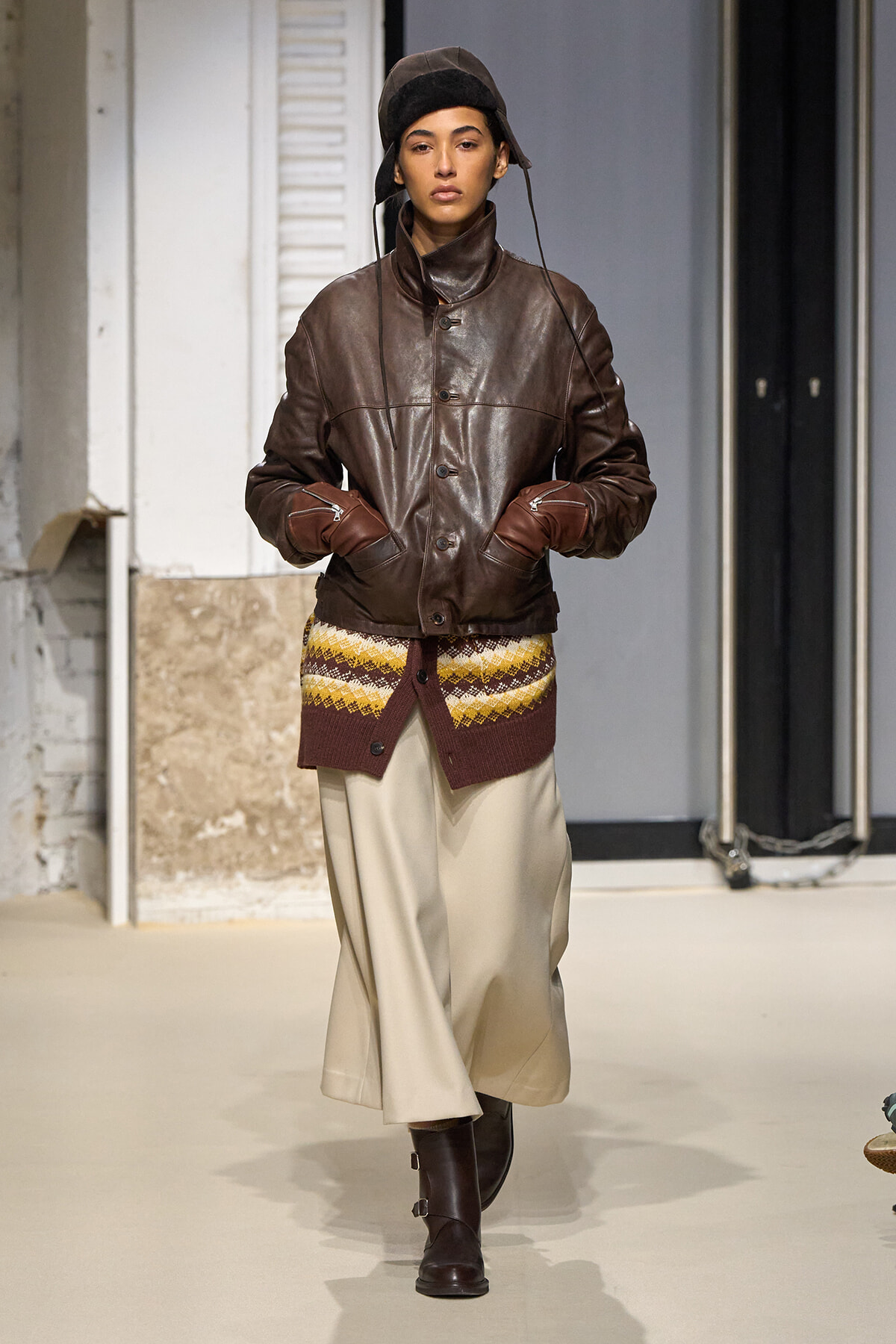 Runway model wearing a brown leather jacket, cream knit layer peeking at the hem, beige wide-leg pants, and dark boots with a black cap.
