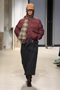 Model walking a runway in a burgundy puffer jacket, plaid scarf, brown cap, dark gray skirt, and brown boots.