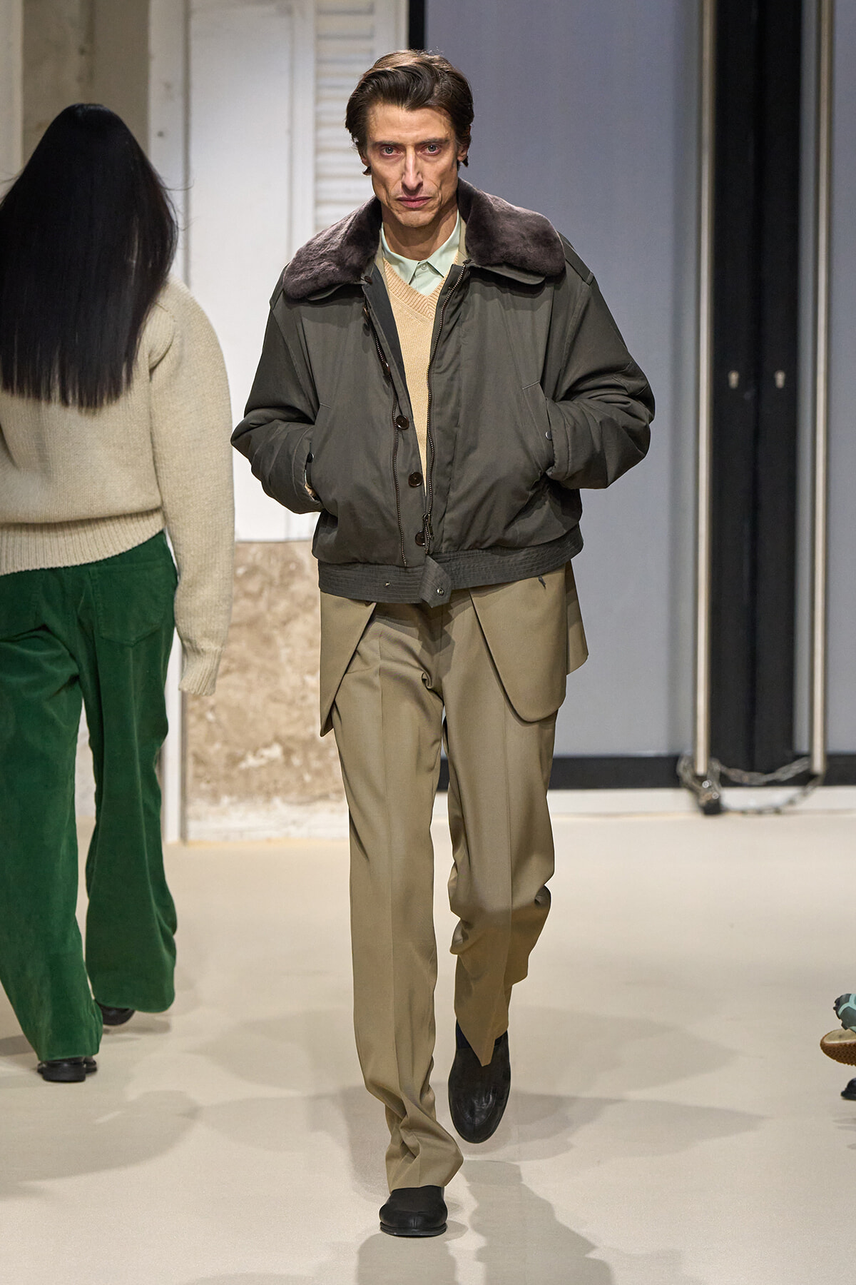 Male model walks toward the camera in an olive bomber with a fur collar, beige sweater, and tan trousers on a runway.