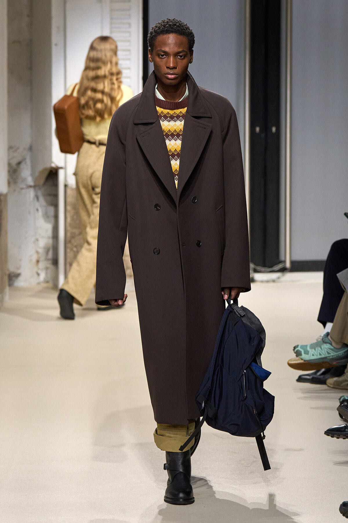 Model walking a runway in a long dark brown double-breasted overcoat over a patterned knit sweater, with khaki cuffs showing, black boots, and a navy backpack held in one hand.