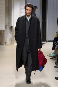 Male fashion model walking the runway in a long dark overcoat with a gray suit, carrying a red backpack with blue details in his right hand.