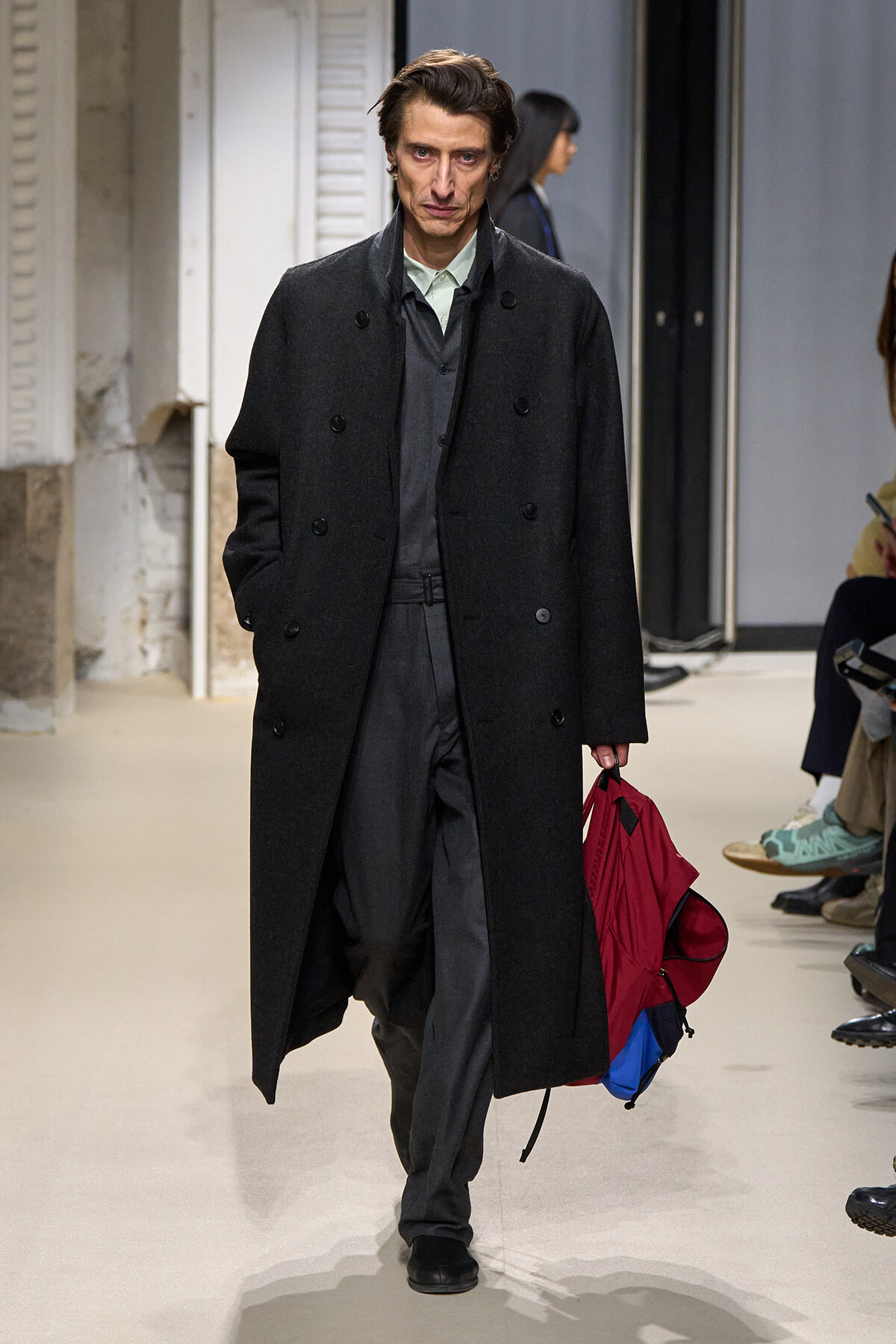 Male fashion model walking the runway in a long dark overcoat with a gray suit, carrying a red backpack with blue details in his right hand.