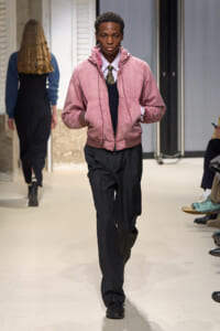 Male model walking a fashion runway in a pink bomber jacket over a white shirt and striped tie, with dark pleated trousers.