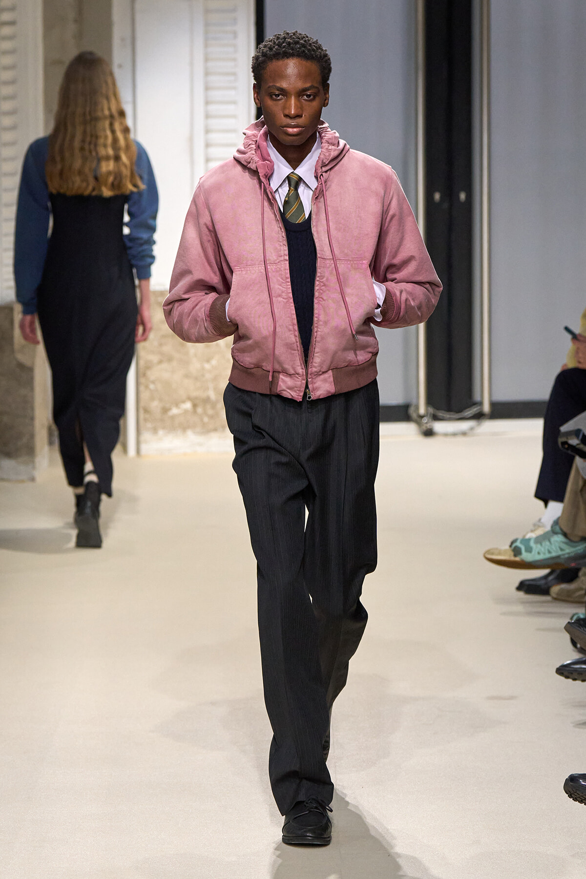 Male model walking a fashion runway in a pink bomber jacket over a white shirt and striped tie, with dark pleated trousers.