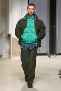 Male model walks the runway wearing an olive-green padded jacket over a bright teal puffer vest and a plaid shirt peeking out, with dark green pants and black shoes.