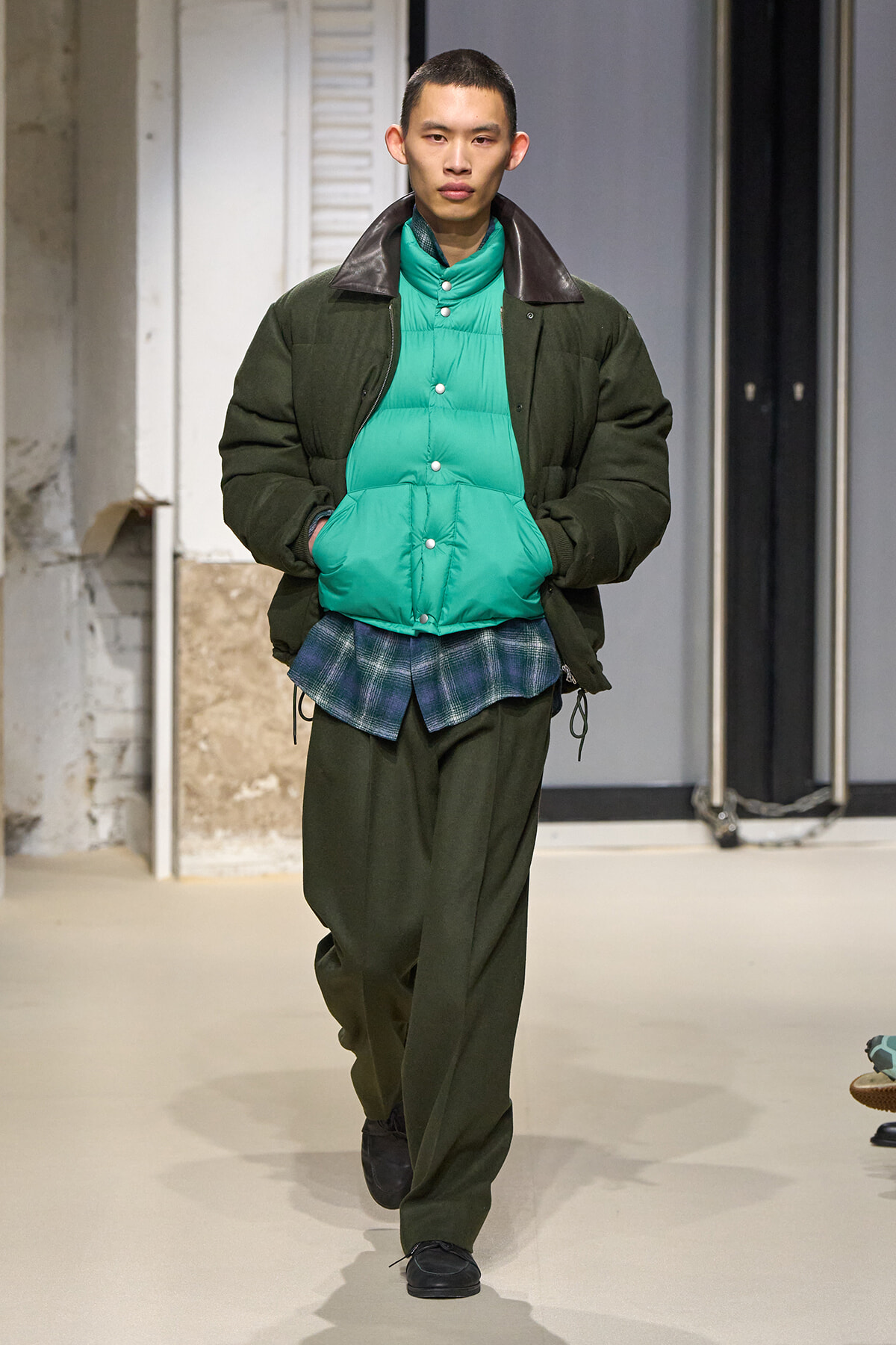 Male model walks the runway wearing an olive-green padded jacket over a bright teal puffer vest and a plaid shirt peeking out, with dark green pants and black shoes.