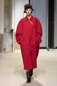 Model on a runway wearing a loose red knee-length coat, black bucket hat, pink crossbody strap, and brown ankle boots with red socks showing at the cuffs.