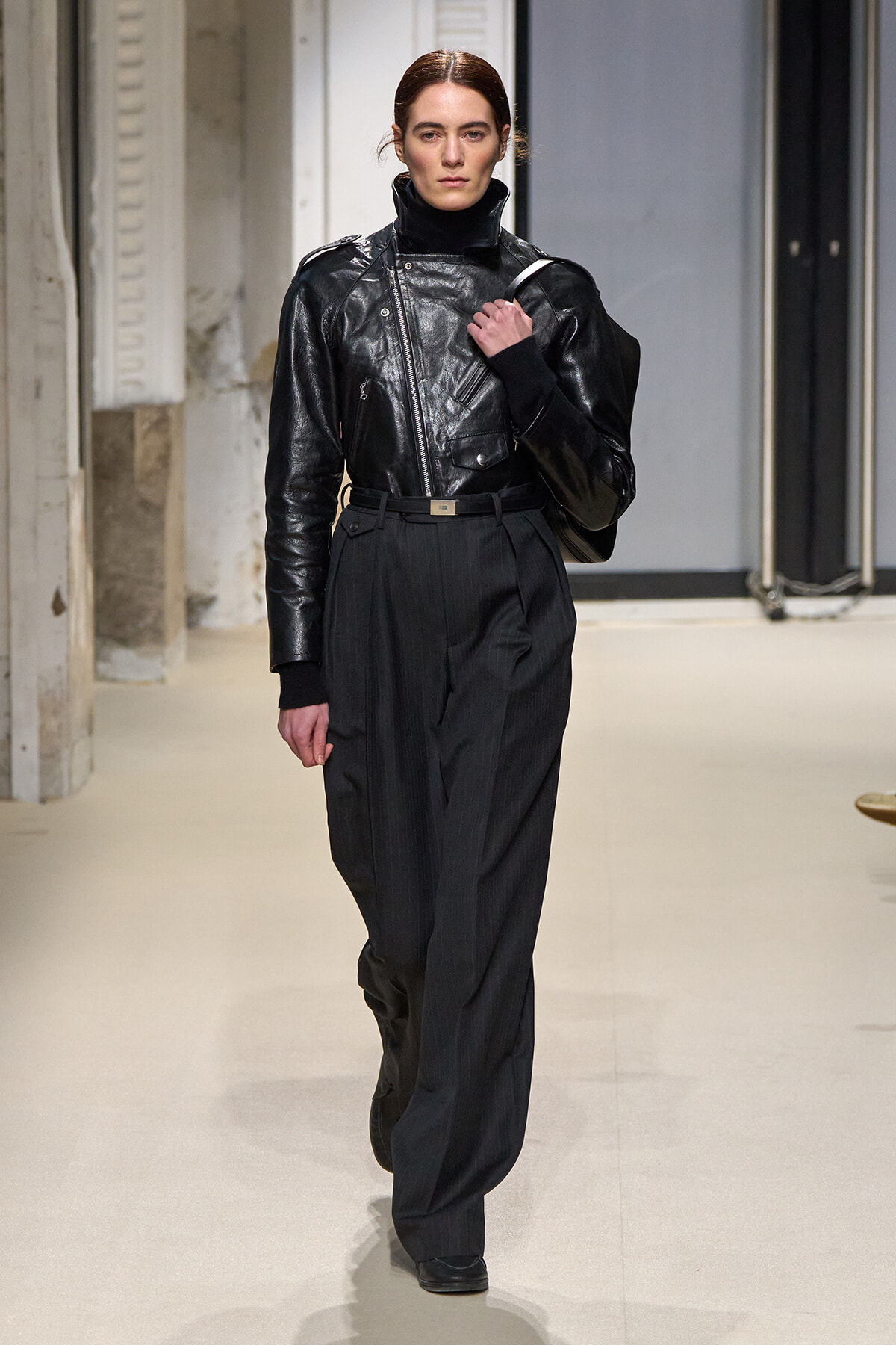 Model walking runway in a black leather jacket, high-waisted wide-leg trousers, and a shoulder bag.