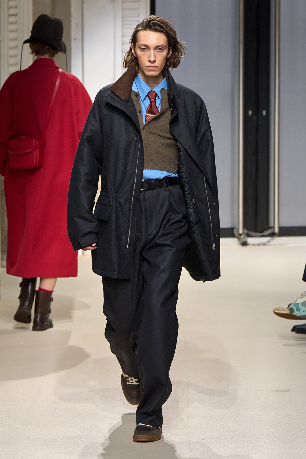 Model walking a runway in a layered menswear look: dark oversized coat, blue shirt, brown vest, red tie, and black trousers with brown shoes.