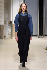 Model walking a runway in a navy blue hoodie layered under a black sleeveless jumpsuit, black boots, neutral backstage setting.