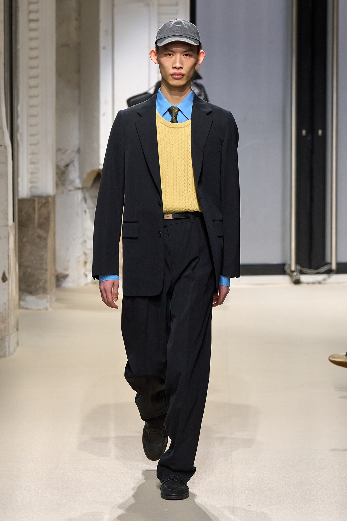 Male model walking a runway in a black suit with a yellow sweater-vest, blue shirt, and striped tie, wearing a gray cap.
