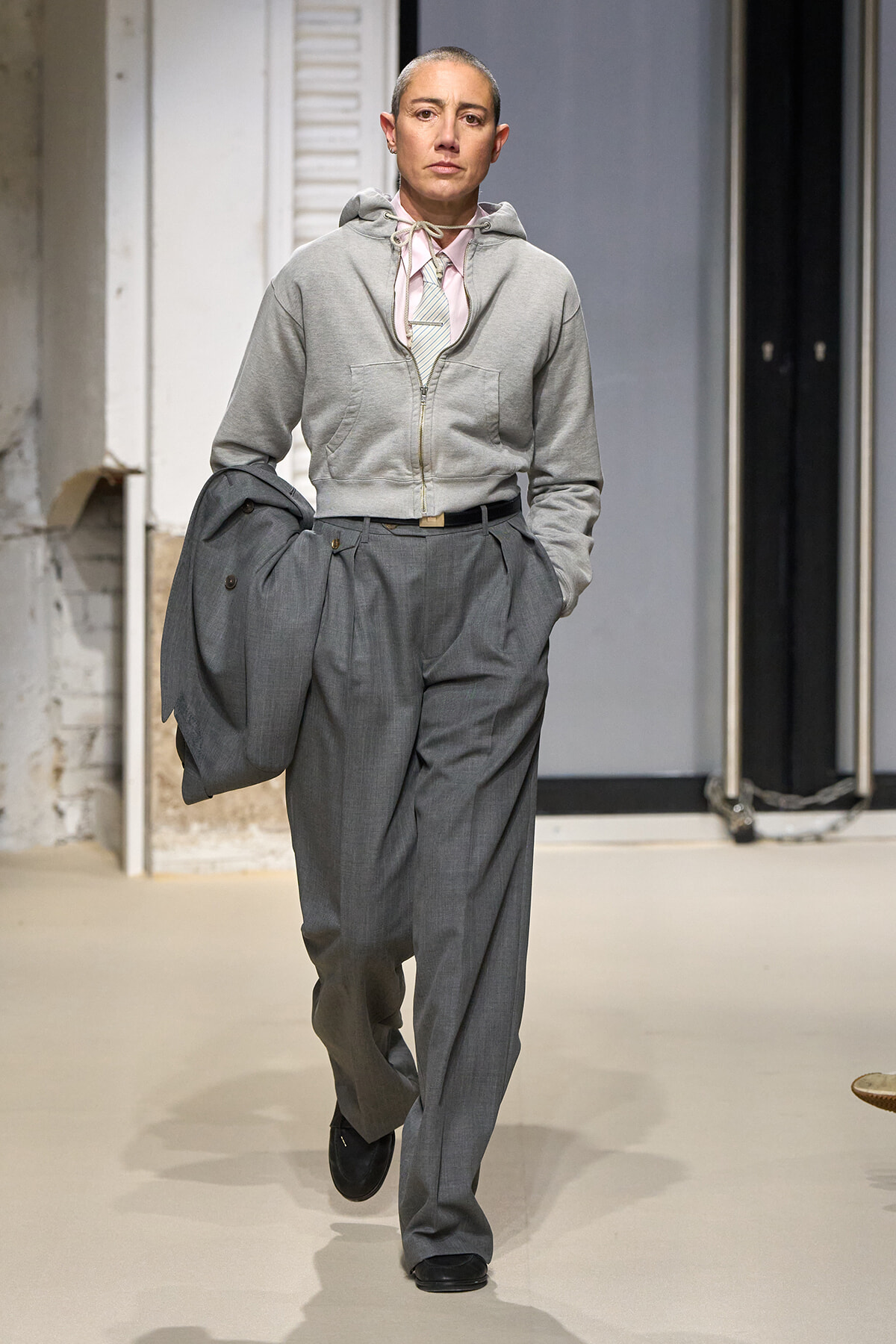 Model walking a runway in oversized gray trousers, a light pink shirt, and a zip hoodie, carrying a gray blazer.