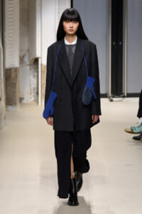 Runway model in a dark pinstripe oversized blazer and wide-leg pants, black boots, with two bright blue knit mittens hanging from a strap across the chest.