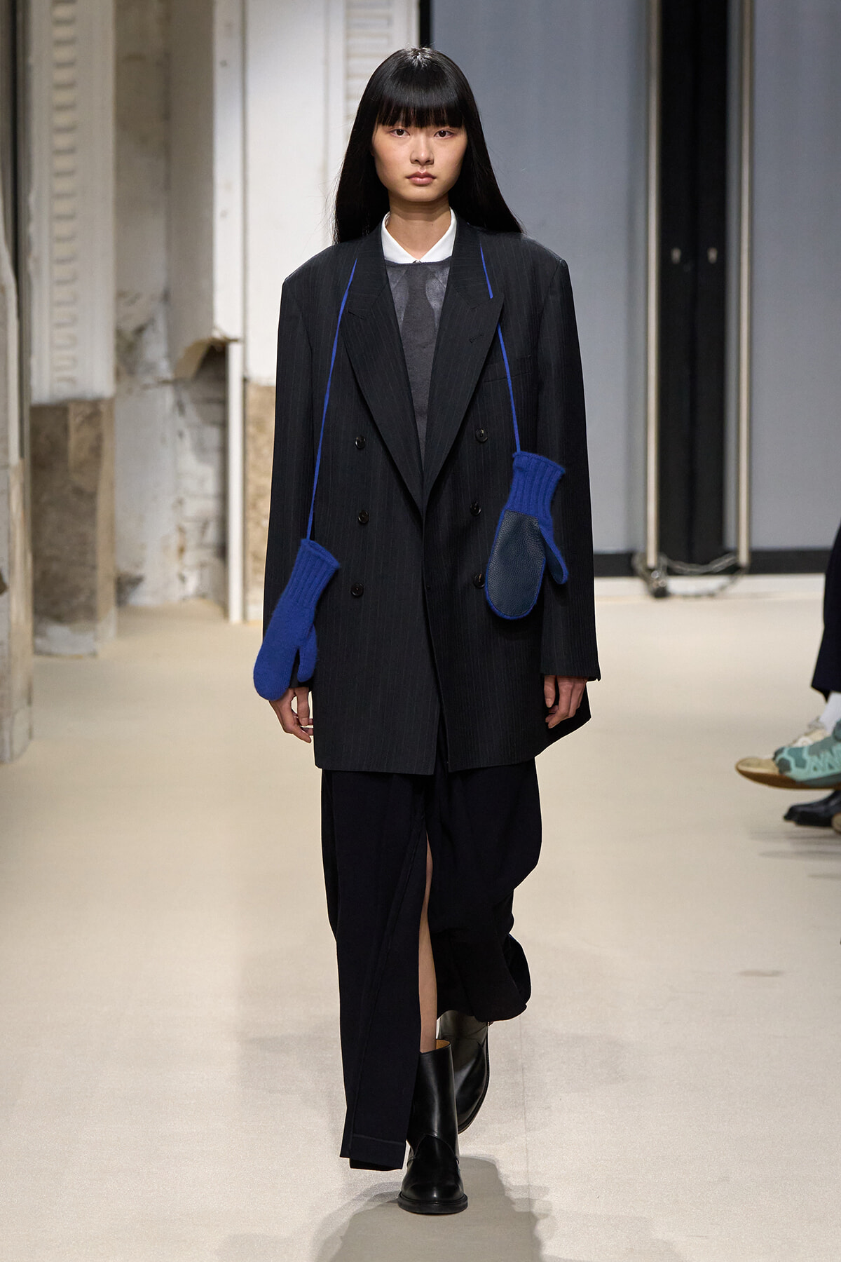Runway model in a dark pinstripe oversized blazer and wide-leg pants, black boots, with two bright blue knit mittens hanging from a strap across the chest.
