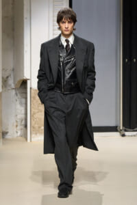 Male model walking the runway in a dark gray suit with a long overcoat and a black leather vest scene-ready ensemble.