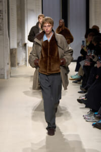Male model walks runway in a gray trench with brown shearling vest, blue shirt, and loose gray trousers with brown gloves.