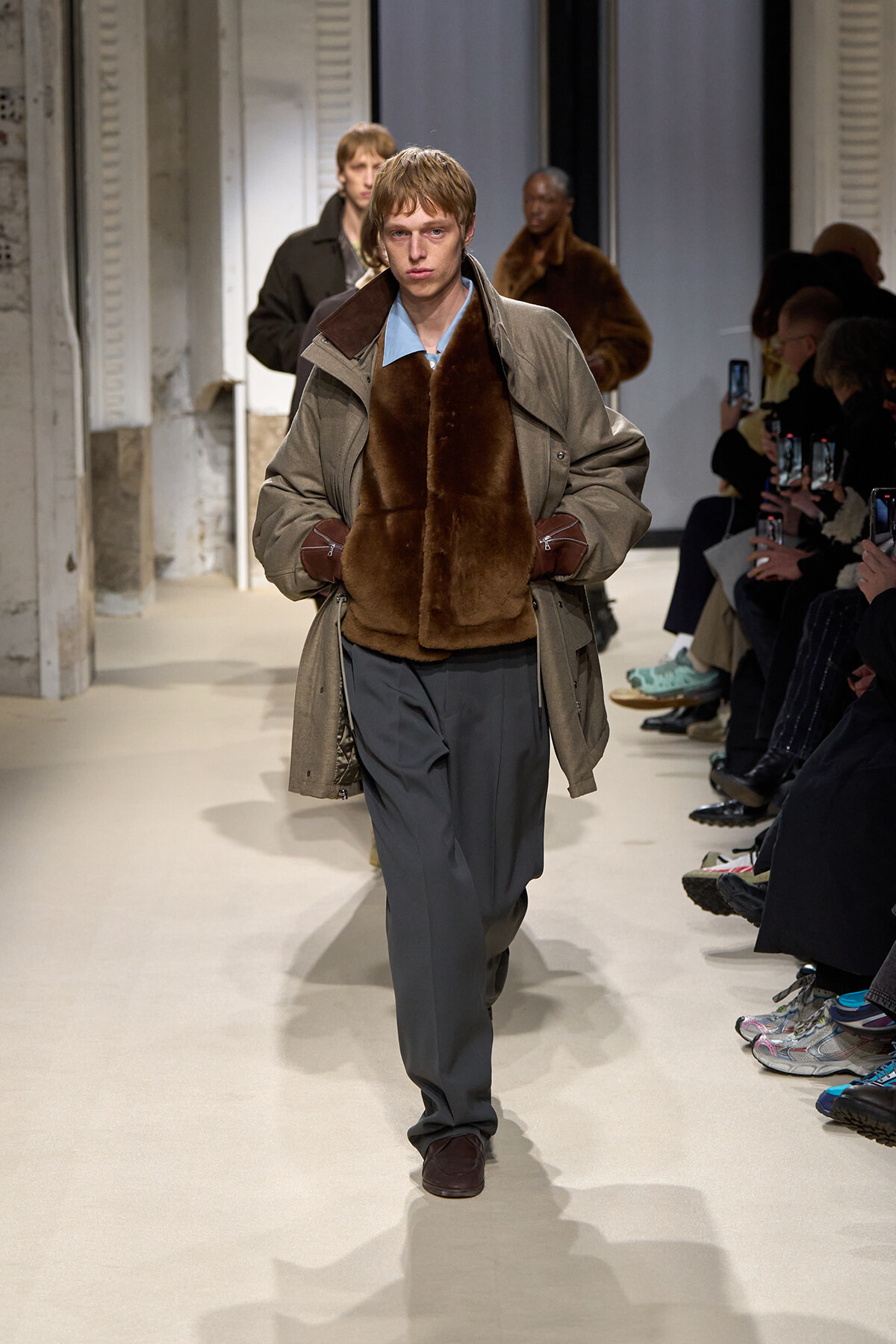 Male model walks runway in a gray trench with brown shearling vest, blue shirt, and loose gray trousers with brown gloves.