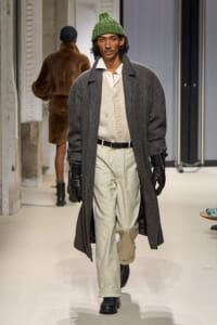 Male model walking runway in oversized dark gray overcoat, cream cardigan, white shirt, beige trousers, black gloves, and a green knit beanie.