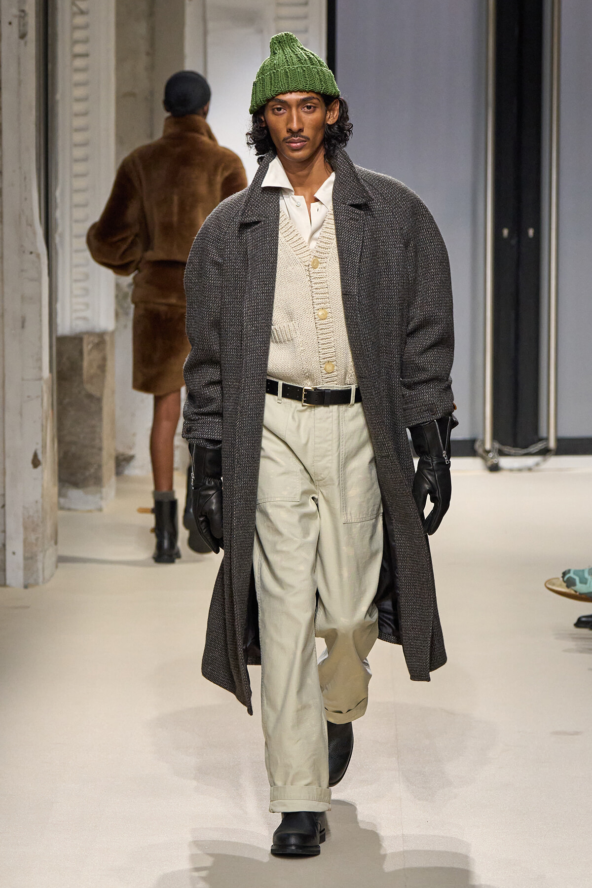 Male model walking runway in oversized dark gray overcoat, cream cardigan, white shirt, beige trousers, black gloves, and a green knit beanie.