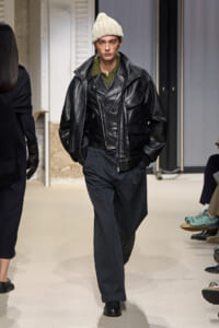 Male model walking the runway in a black leather oversized jacket over a green knit sweater, dark wide‑leg pants, and a cream cable-knit beanie.