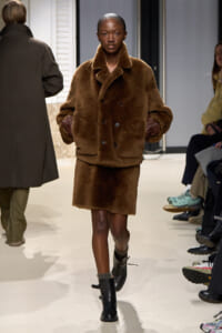 Model walks a fashion runway in a oversized brown fur coat with a wide collar and matching fur-trimmed short skirt, black ankle boots.