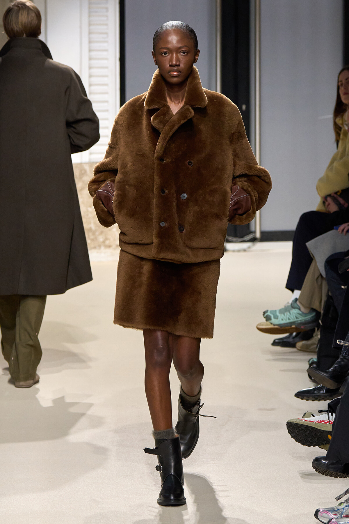 Model walks a fashion runway in a oversized brown fur coat with a wide collar and matching fur-trimmed short skirt, black ankle boots.