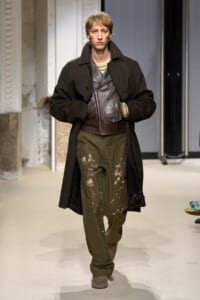 Male model walks the runway in a long dark coat over a brown leather zippered vest and olive cargo pants splattered with paint, beige striped knit underlayer, and brown shoes.