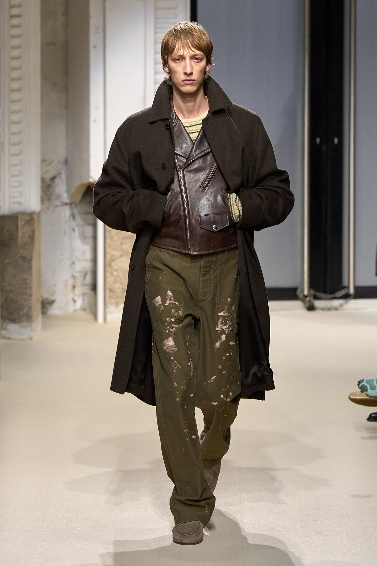 Male model walks the runway in a long dark coat over a brown leather zippered vest and olive cargo pants splattered with paint, beige striped knit underlayer, and brown shoes.