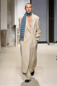 Model on a runway wearing a long beige double-breasted coat with a blue plaid scarf draped around the neck.