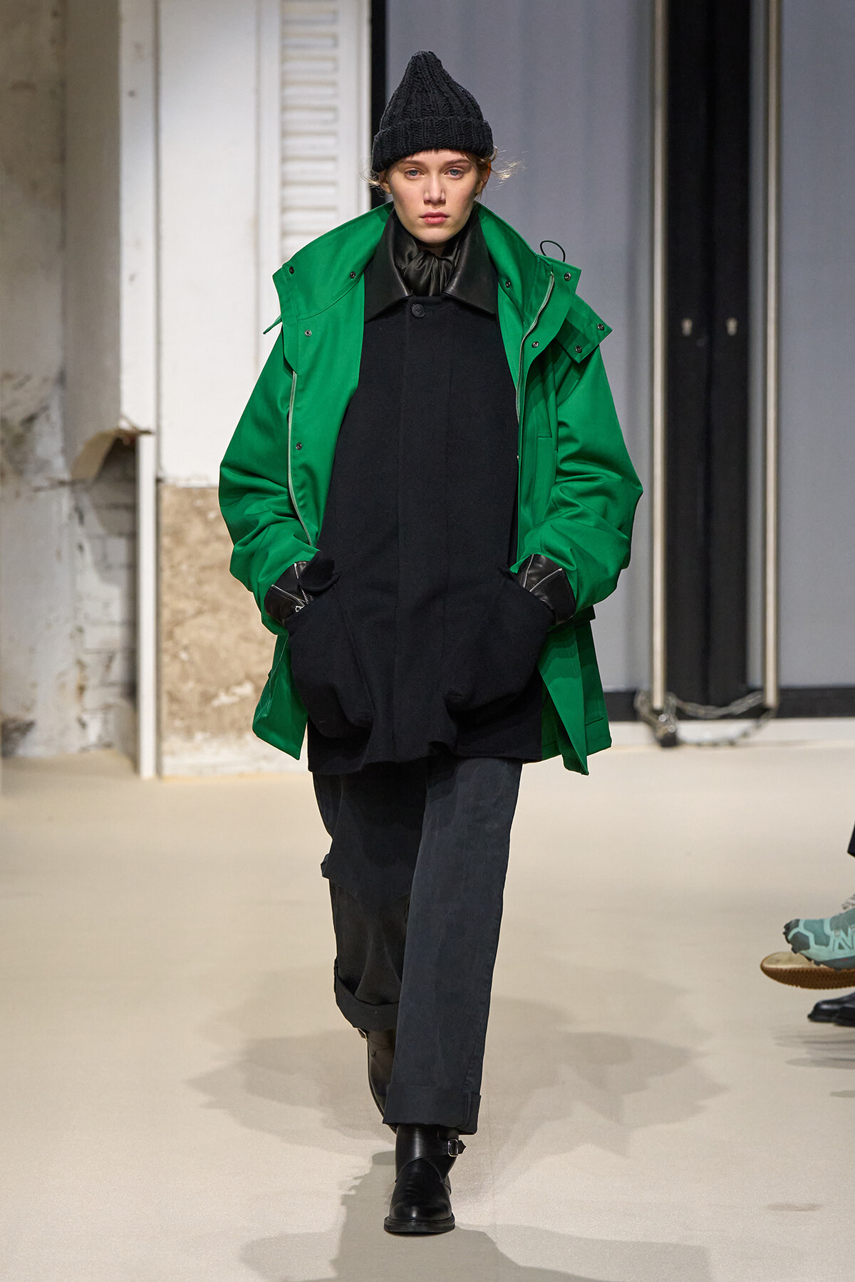 Model walking a fashion show wearing a bright green parka over a loose black tunic, black beanie and gloves.