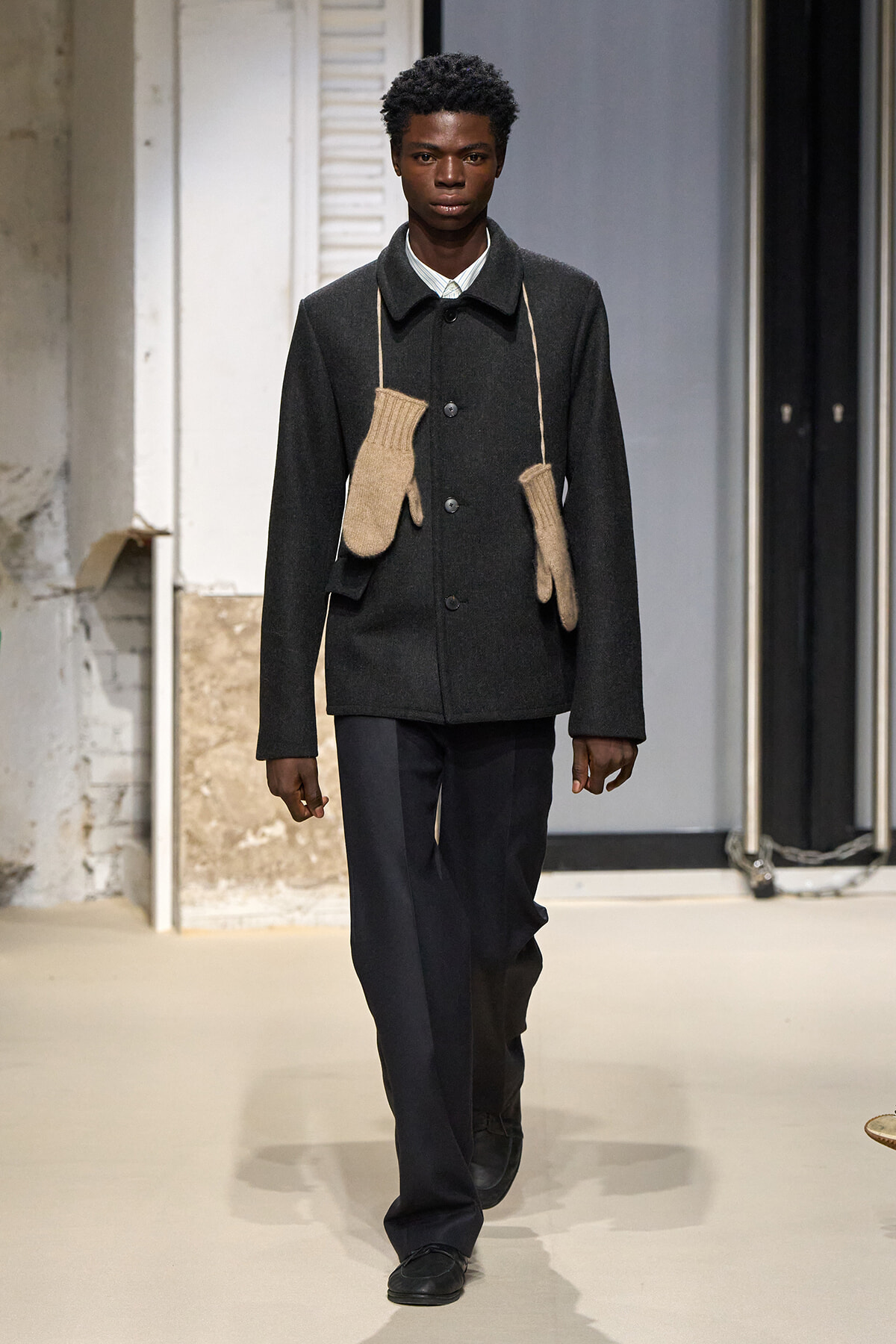 Male model walks a runway wearing a dark gray wool coat with beige knit mittens hanging from cords over his shoulders.",