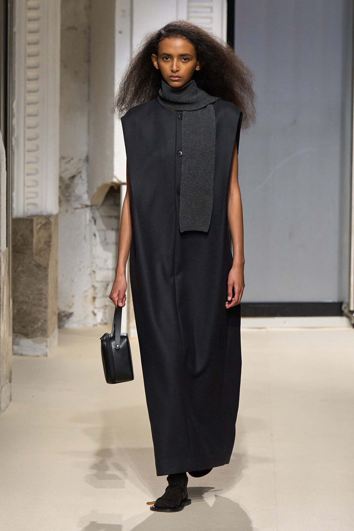 Female model walking a runway in a long black sleeveless coat with a gray scarf and small black handbag.