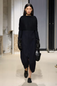 Runway model in a navy dress with a wide black cape-like top, black gloves, and a small black handbag.