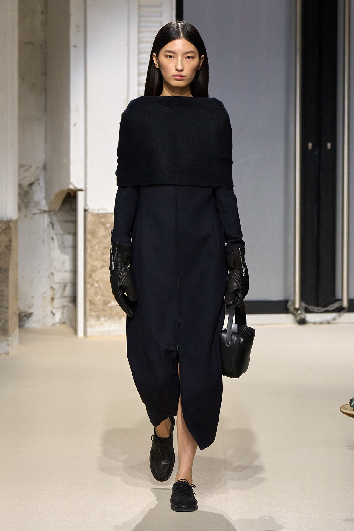 Runway model in a navy dress with a wide black cape-like top, black gloves, and a small black handbag.