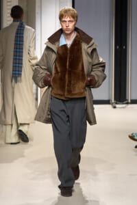 Male model walks a runway in a brown fur vest over a tan parka and blue shirt, with gray wide-leg trousers and brown gloves.