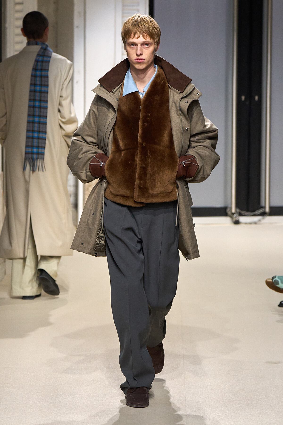 Male model walks a runway in a brown fur vest over a tan parka and blue shirt, with gray wide-leg trousers and brown gloves.