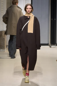 Model walking a runway in a long dark brown coat, beige scarf draped around the neck, white crossbody strap, and cream sneakers with red-and-white socks.