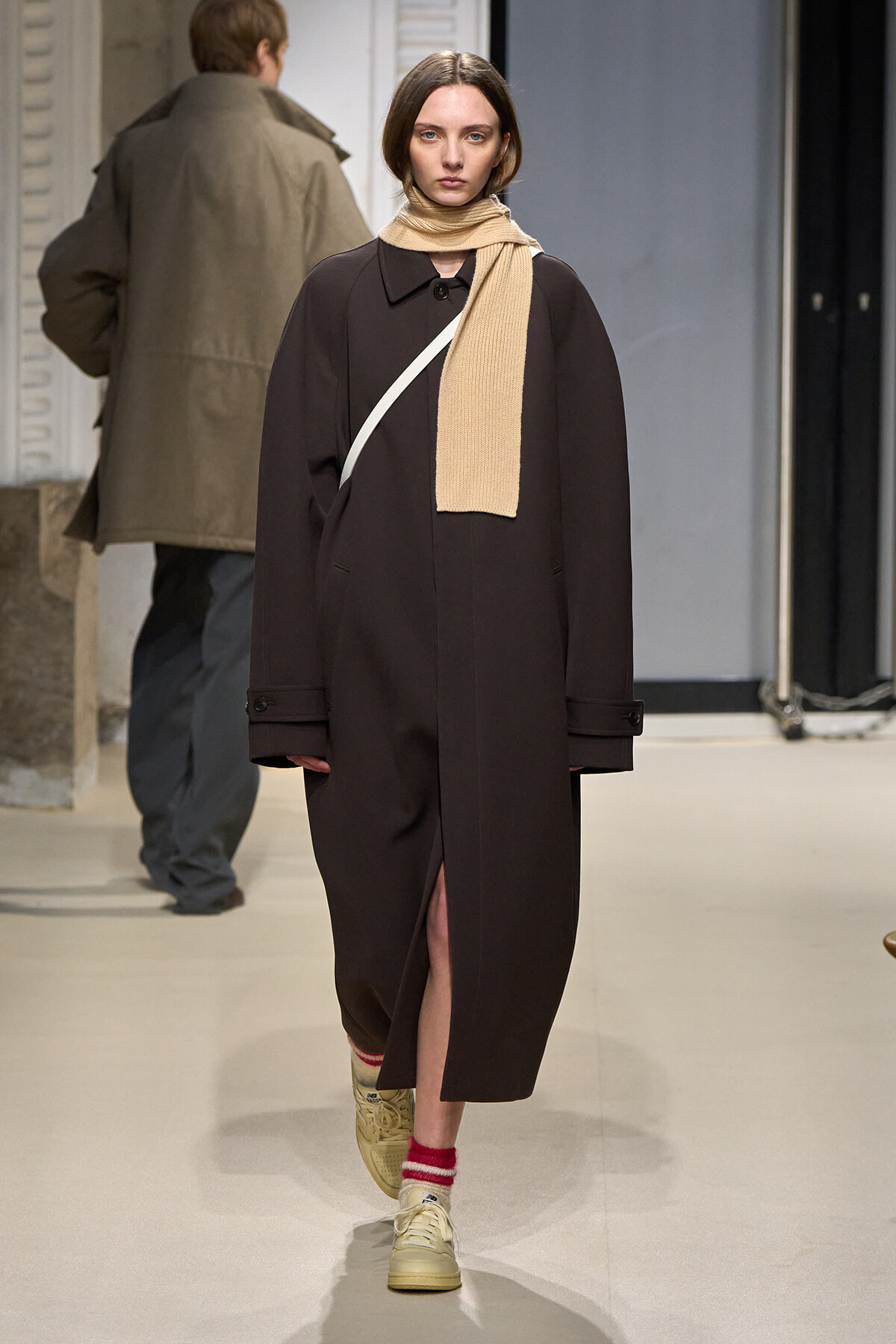 Model walking a runway in a long dark brown coat, beige scarf draped around the neck, white crossbody strap, and cream sneakers with red-and-white socks.