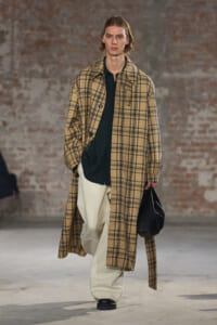 Model on a runway wearing a long tan plaid coat over a black shirt and oversized cream trousers, carrying a black bag.